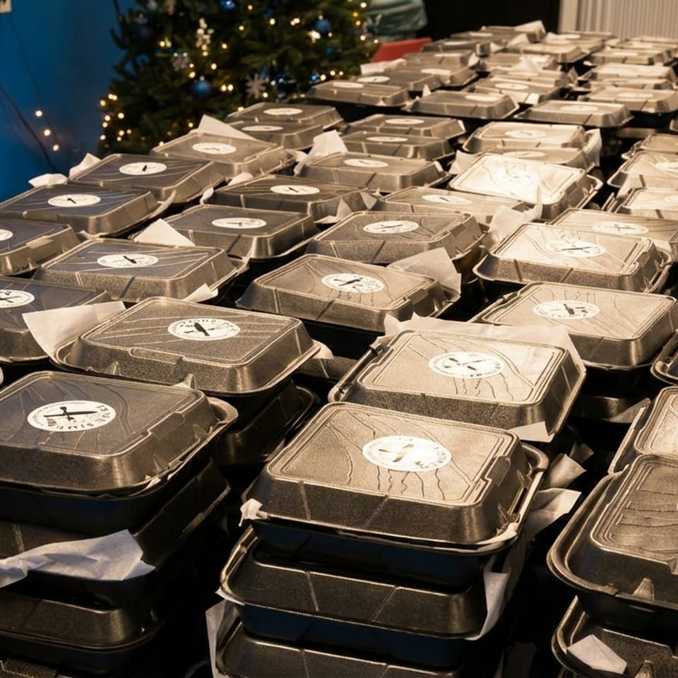 Numerous black takeout containers with white circular stickers are stacked on a table, ready for distribution.