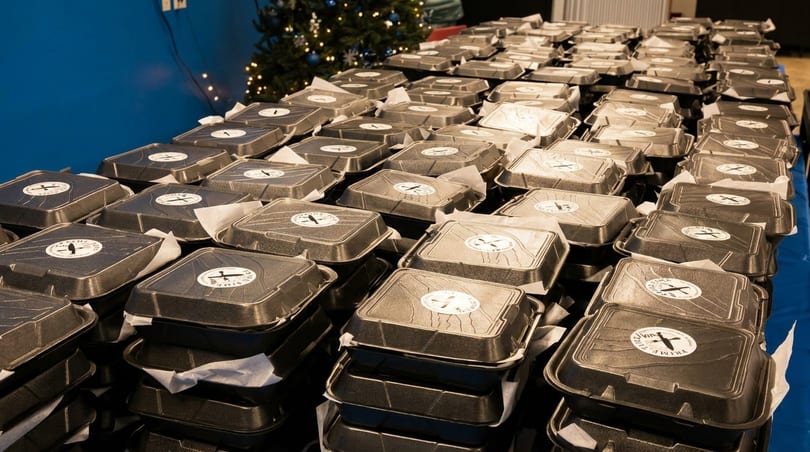 Numerous black takeout containers with white circular stickers are stacked on a table, ready for distribution.