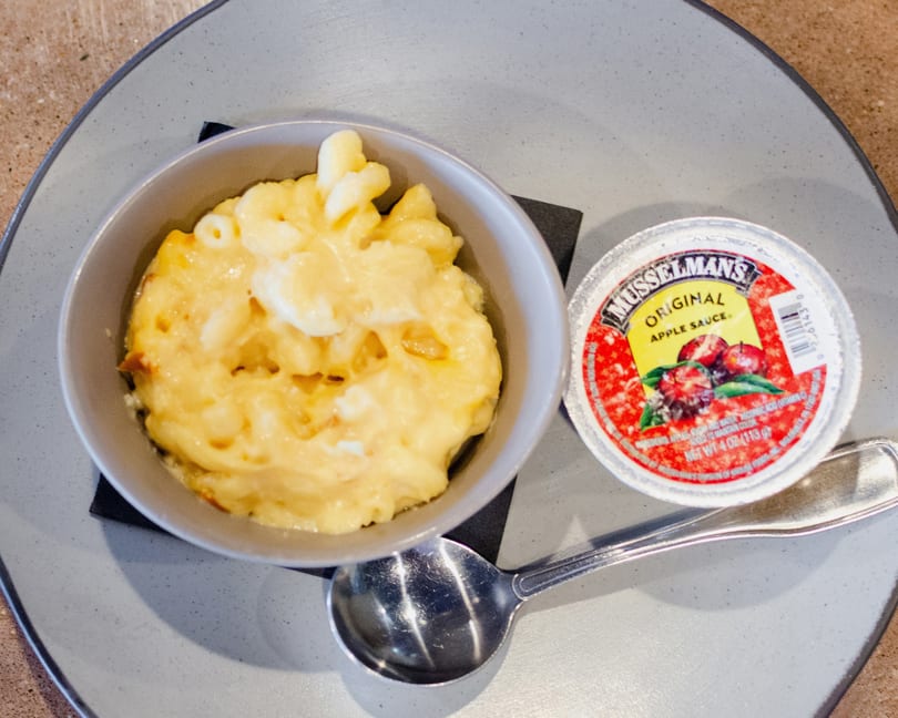 A bowl of macaroni and cheese with a side of Musselman's Original Apple Sauce and a spoon.
