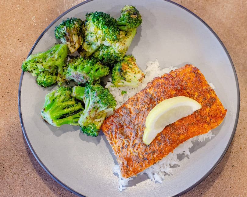A plate of seasoned salmon with a lemon wedge, white rice, and roasted broccoli.