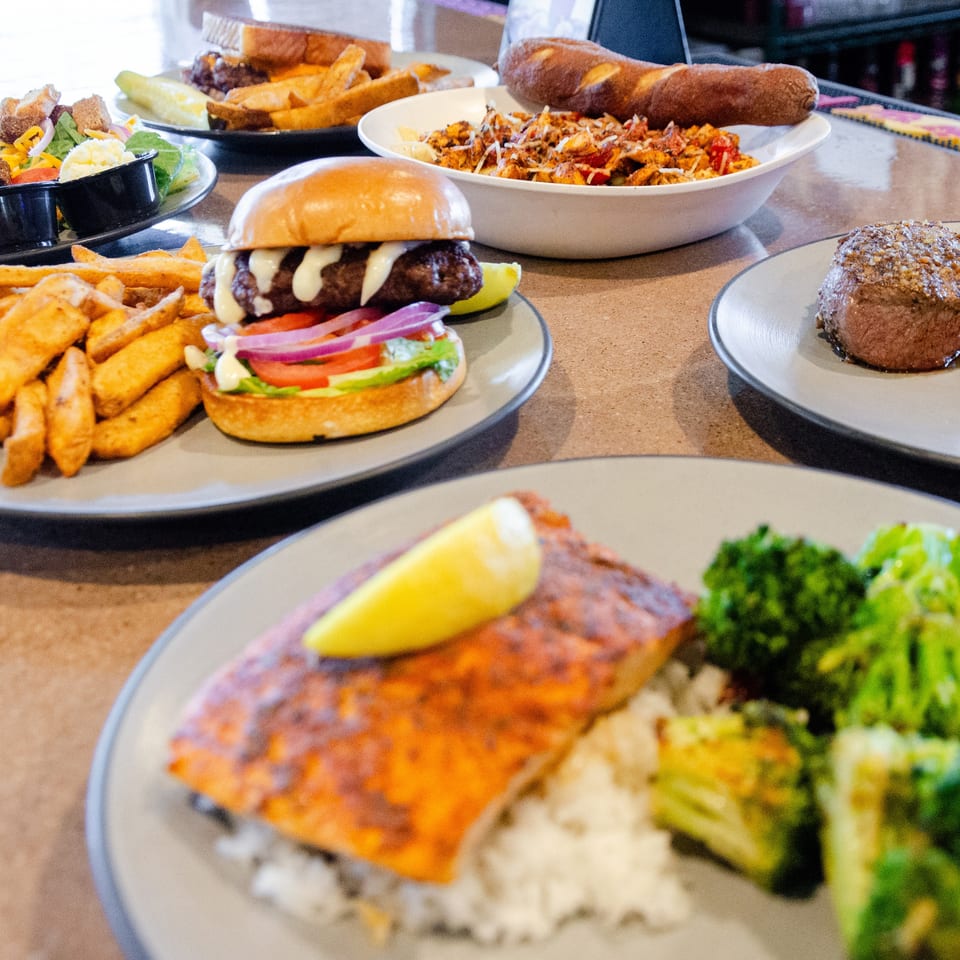 A table laden with various dishes, including salmon, steak, a burger with fries, and pasta.