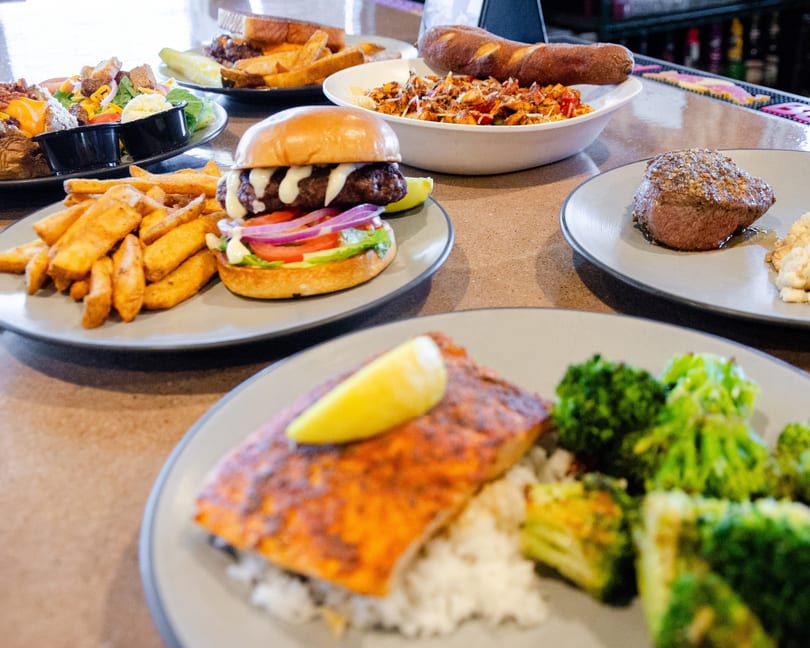 A table laden with various dishes, including salmon, steak, a burger with fries, and pasta.