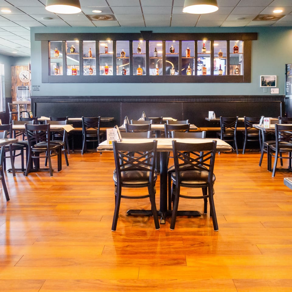 An empty restaurant dining room with wooden floors, black tables, and a wall display of liquor bottles.