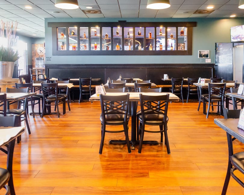 An empty restaurant dining room with wooden floors, black tables, and a wall display of liquor bottles.