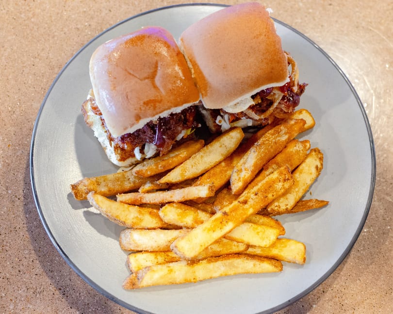 Two sliders with pulled pork and coleslaw, served with a side of seasoned french fries on a gray plate.