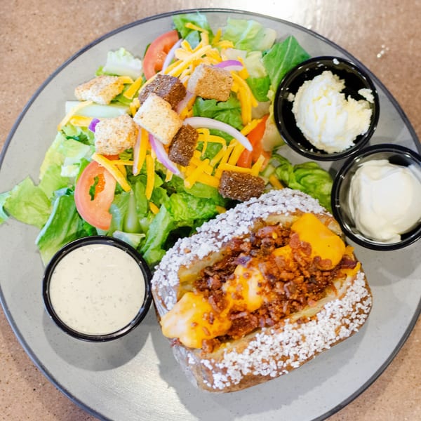 Loaded baked potato with bacon and cheese, a side salad, butter, sour cream, and ranch dressing.
