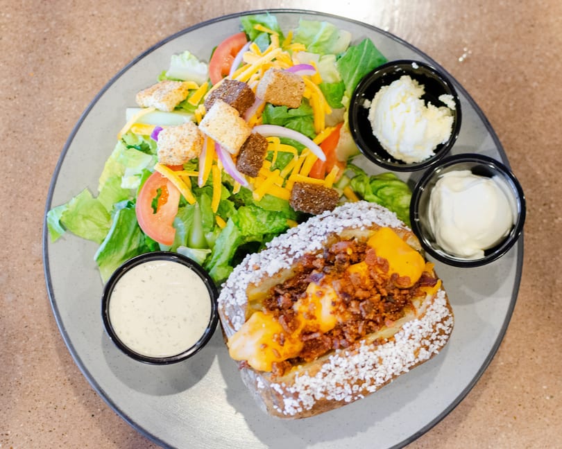 Loaded baked potato with bacon and cheese, a side salad, butter, sour cream, and ranch dressing.