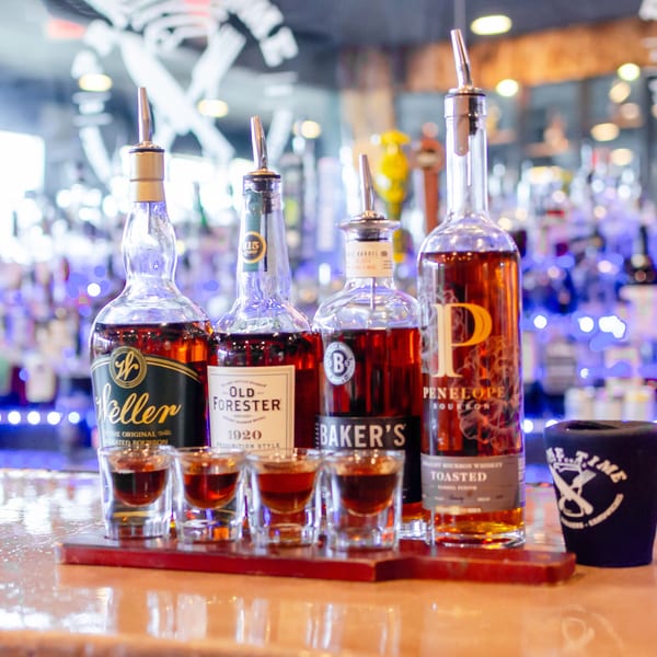 Four bottles of bourbon with shot glasses filled with whiskey on a wooden tray at a bar.