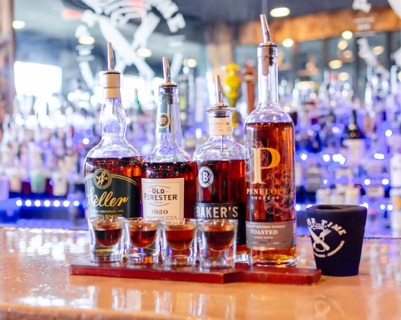 Four bottles of bourbon with shot glasses filled with whiskey on a wooden tray at a bar.