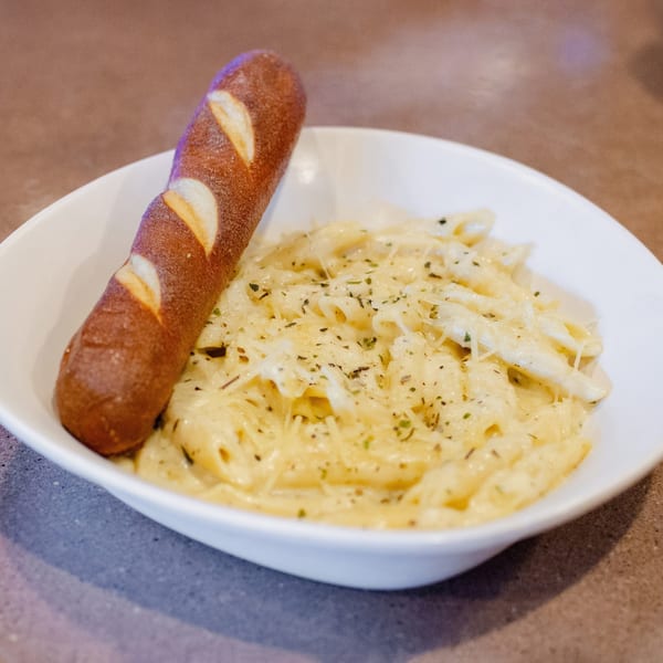 Creamy penne pasta with herbs and cheese, served with a pretzel breadstick in a white bowl.