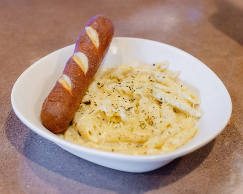 Creamy penne pasta with herbs and cheese, served with a pretzel breadstick in a white bowl.
