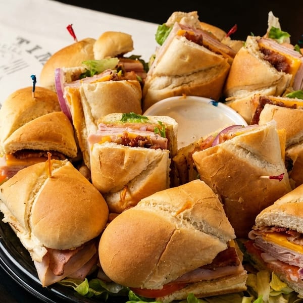 A black platter filled with various Prime Time Deli sandwiches and sliders, with a side of dipping sauce.