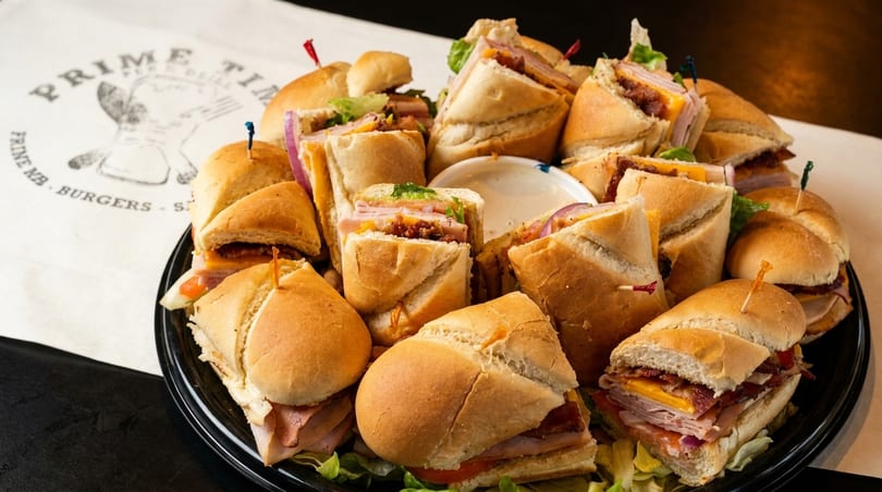 A black platter filled with various Prime Time Deli sandwiches and sliders, with a side of dipping sauce.