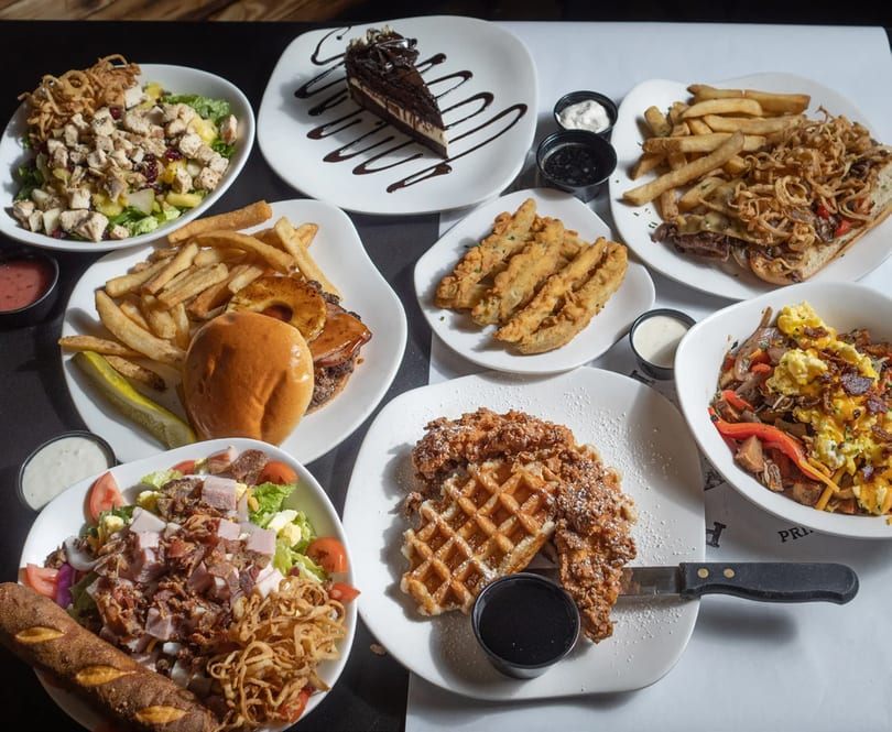 A table laden with various dishes including salads, burgers, fries, fried pickles, and dessert.