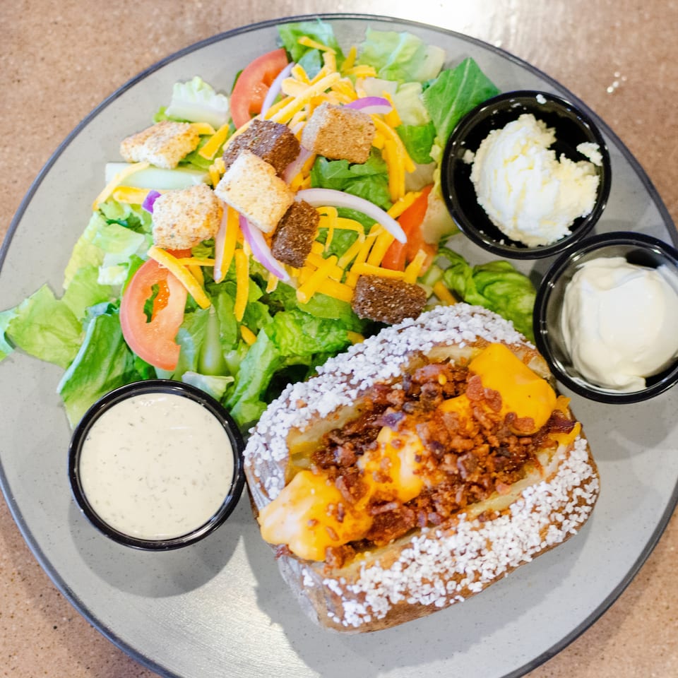 Loaded baked potato with bacon and cheese, a side salad, butter, sour cream, and ranch dressing.