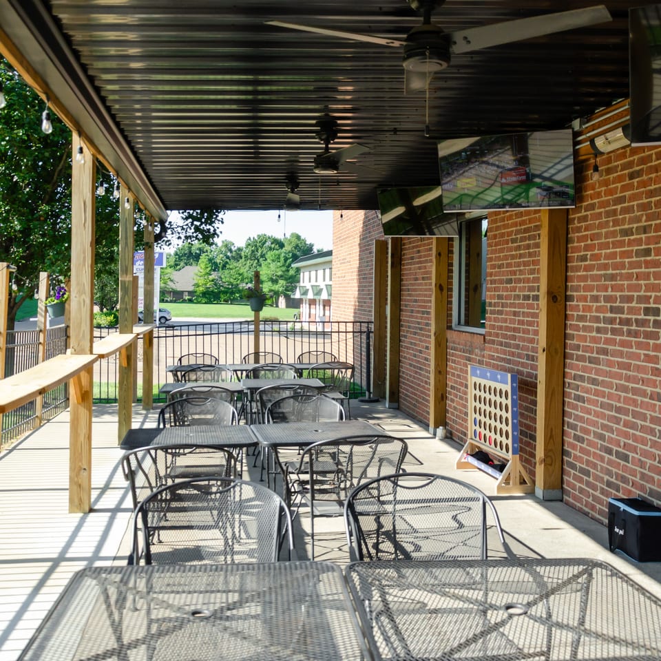 Outdoor patio with metal tables and chairs, brick wall, string lights, ceiling fans, and TVs.