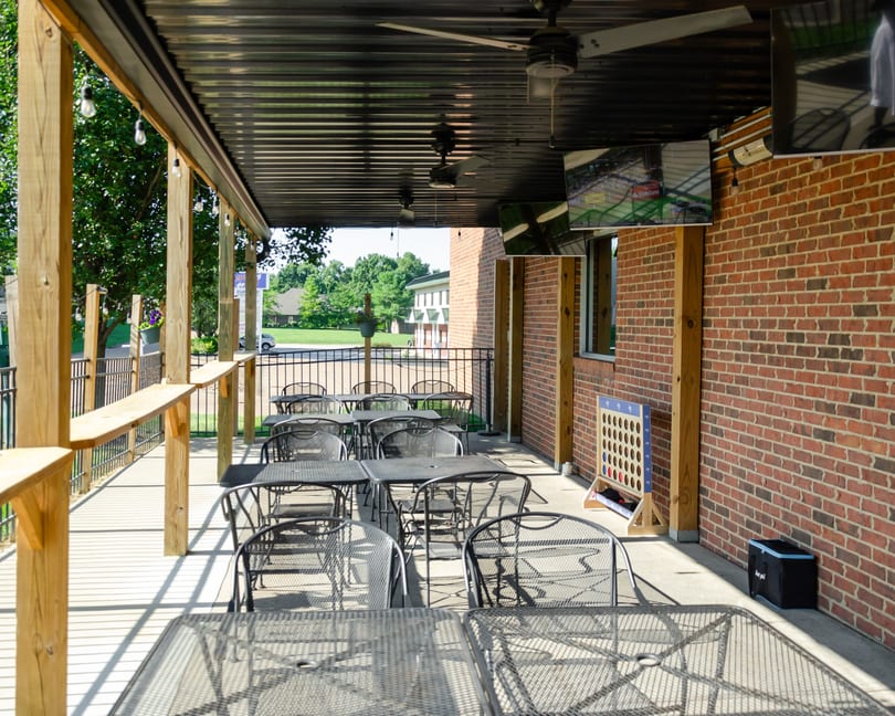 Outdoor patio with metal tables and chairs, brick wall, string lights, ceiling fans, and TVs.