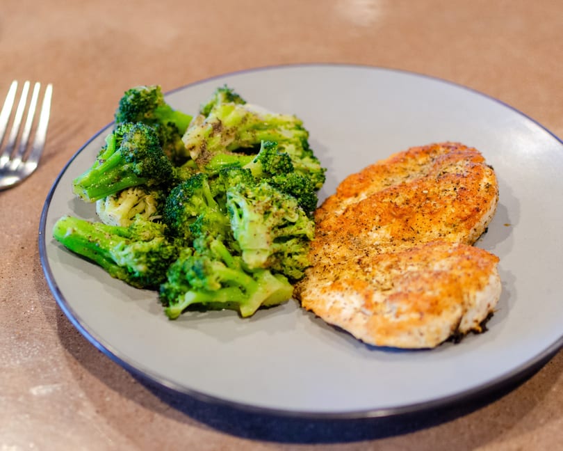 Seasoned chicken breast and steamed broccoli florets on a gray plate with a fork nearby.