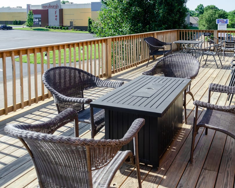 Outdoor dining deck with dark wicker chairs and tables, overlooking a parking lot and distant buildings.