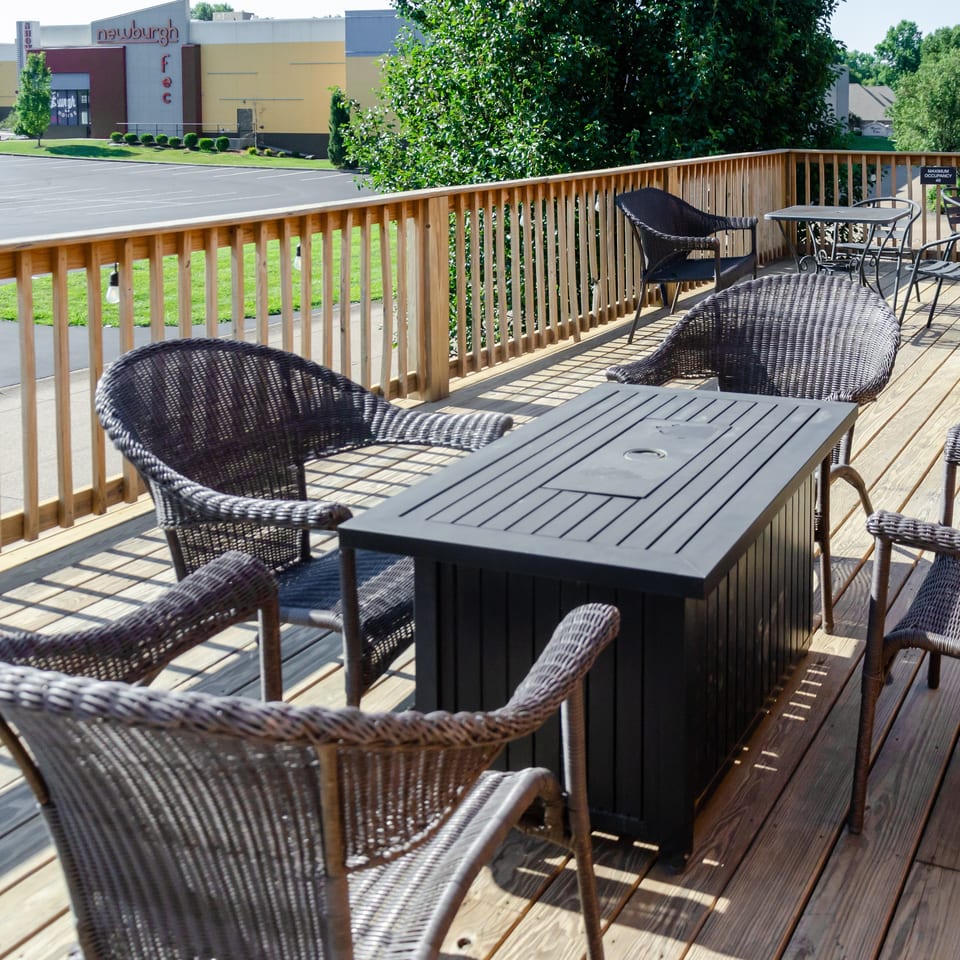 Outdoor dining deck with wicker chairs and dark tables, overlooking a parking lot and distant buildings.