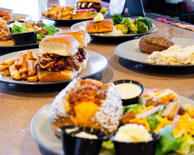 A table filled with various dishes including sliders, steak, salmon, salads, and fries.
